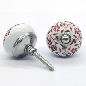Op de afbeelding: Twee witte keramische kastknoppen met een rood bloemenpatroon. Eén knop heeft een zilveren schroef en de andere een zilveren basis.