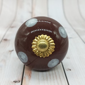 Puede incluir: Un pomo de cerámica marrón con lunares blancos y un acento de metal dorado. El pomo es redondo y tiene una superficie lisa.