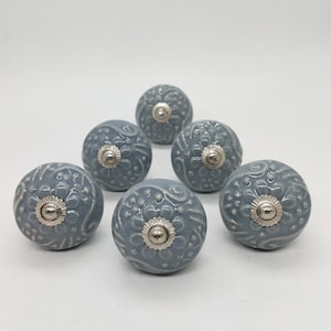 May include: Six grey ceramic cabinet knobs with a silver metal accent in the centre. The knobs have a floral design.