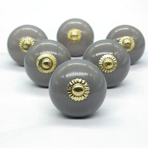 Könnte beinhalten: Sechs graue Keramik-Schrankknöpfe mit goldenen Akzenten. Jeder Knopf hat ein kreisförmiges Design mit einem erhöhten, geriffelten Rand.