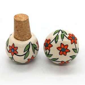 Può includere: Un set di due tappi per bottiglie in ceramica bianca con un motivo floreale. I tappi presentano fiori arancioni con foglie verdi e centri blu. Un tappo ha un tappo di sughero.