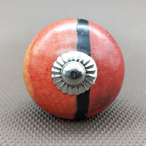 May include: A round, red and black cabinet knob with a silver metal center. The knob has a black stripe and a silver metal center with a decorative design.
