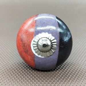 Op de afbeelding: Een ronde keramische knop met een zilveren metalen accent. De knop is in drie kleuren geschilderd: rood, paars en zwart.
