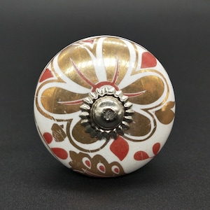 Op de afbeelding: Witte keramische knop met een gouden en rood bloemenmotief. De knop heeft een zilveren metalen basis.