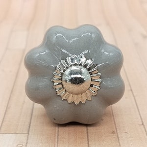 以下が含まれることがあります： 中央に銀色の花の形をしたアクセントが付いた灰色のセラミック製のキャビネットノブです。