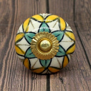 Könnte beinhalten: Ein Keramik-Knopf mit weißem Hintergrund und einem geometrischen Muster in Gelb und Türkis. Der Knopf hat eine goldene Metallmitte.