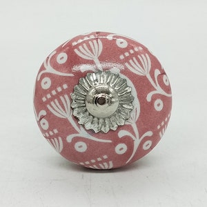 Op de afbeelding: Een ronde, roze keramische kastknop met een wit bloemmotief. De knop heeft een zilverkleurige metalen basis. Deze decoratieve hardware is geschikt voor laden, kasten en meubels.