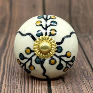 Könnte beinhalten: Weißer Keramik-Knopf mit einem schwarzen und grünen Blumenmuster und einer goldenen Mitte.