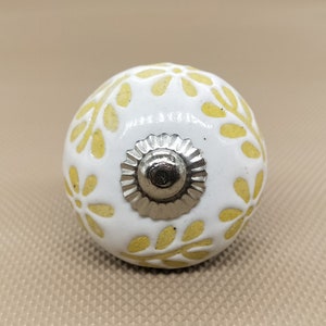 以下が含まれることがあります： 白いセラミックキャビネットノブ、黄色の花柄。ノブは丸い形で、銀色の金属ベースが付いています。この装飾的なハードウェアは、引き出しやキャビネットに適しています。