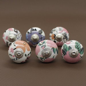 Puede incluir: Seis pomos de cajón de cerámica con patrones florales en tonos de rosa, morado y verde. Cada pomo tiene una base de metal plateado.