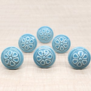 Peut inclure: Six boutons de meuble en céramique bleu clair avec un motif floral. Les boutons sont ronds avec un motif de fleur en relief au centre et une bordure circulaire. Les boutons sont disposés sur une surface claire imitation bois.