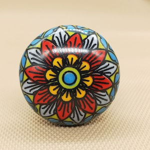 May include: A round ceramic knob with a colorful floral design. The flower has red petals, a yellow center, and a blue circle in the middle. The background is a black and white geometric pattern.