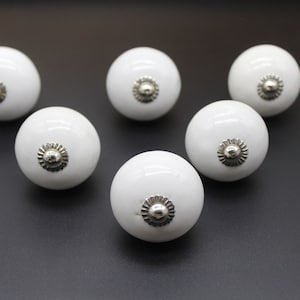 May include: Six white ceramic cabinet knobs with a glossy finish and silver-colored metal accents. These round knobs are arranged on a dark surface, suitable for furniture hardware and home decor projects.