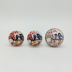 May include: Three round ceramic knobs with a white background and a floral design in shades of red, pink, and gold. Each knob features two blue birds perched on a branch.