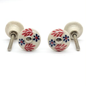May include: Two white ceramic drawer knobs with red and blue floral designs. The knobs have a silver screw.