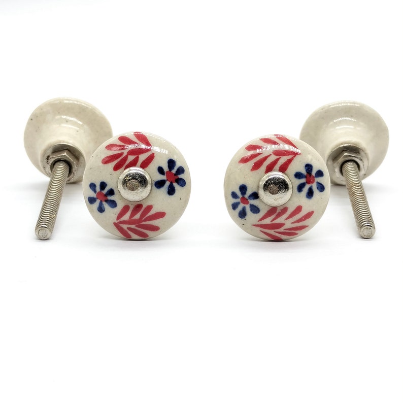 May include: Two white ceramic drawer knobs with red and blue floral designs. The knobs have a silver screw.