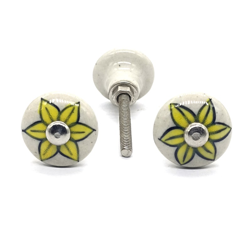 K&ouml;nnte beinhalten: Zwei wei&szlig;e Keramik-Schubladengriffe mit gelben Blumendesigns. Die Griffe haben eine silberne Schraube.