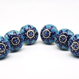 Op de afbeelding: Zes blauwe en witte keramische kastknoppen met een stervormig ontwerp. De knoppen hebben een wit midden met blauwe stippen en een blauw stervormig patroon rond het midden.