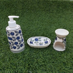 May include: A white ceramic bathroom set with a blue floral design. The set includes a soap dispenser, a soap dish, and an oil burner.