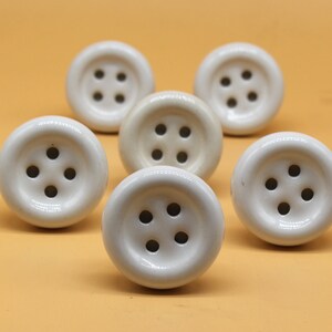 May include: Six white ceramic buttons with four holes each. The buttons are arranged in a semi-circle on a light orange surface.