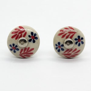 May include: Two white ceramic drawer knobs with red and blue floral designs. The knobs have a silver metal center.
