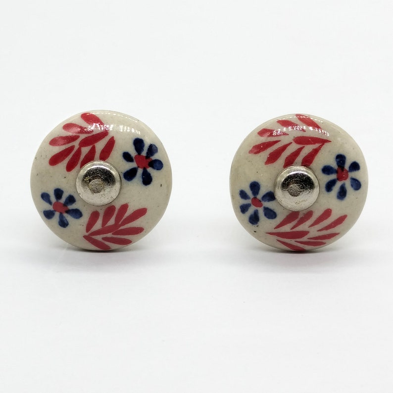 May include: Two white ceramic drawer knobs with red and blue floral designs. The knobs have a silver metal center.