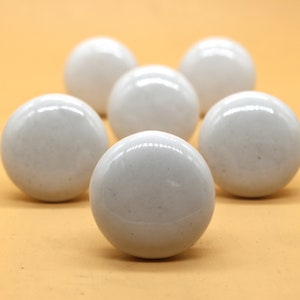 May include: Six white ceramic knobs with a slightly textured surface. The knobs are round and have a smooth, glossy finish.