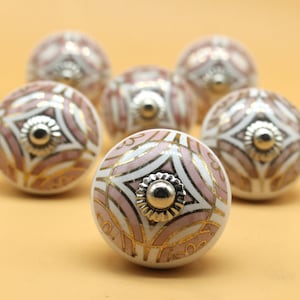 Op de afbeelding: Zes witte keramische kastknoppen met een geometrisch ontwerp in roze en goud. Elke knop heeft een zilveren metalen midden.