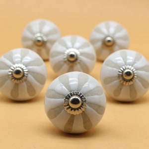 May include: Six white ceramic cabinet knobs with a raised, scalloped design and a silver metal center. The knobs have a subtle beige stripe pattern.