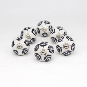 Op de afbeelding: Acht witte keramische knoppen met een zwart bladmotief. De knoppen hebben een zilveren metalen accent in het midden.