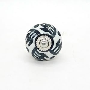 Könnte beinhalten: Ein weißer Keramik-Knopf mit einem schwarzen Blumenmuster und einer silbernen Metallmitte.