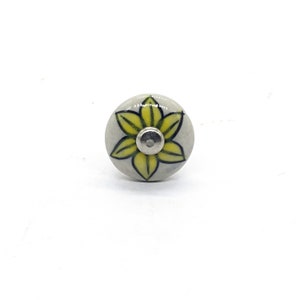 May include: A white ceramic knob with a yellow flower design. The flower has five petals and a black outline. The knob has a silver center.