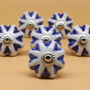 以下が含まれることがあります： 青い花柄の白いセラミック製のキャビネットノブが6個。ノブの中央は銀色の金属製です。
