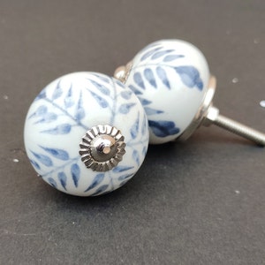 Könnte beinhalten: Zwei weiße Keramik-Schrankknöpfe mit einem blauen Blumenmuster. Die Knöpfe haben eine silberne Metallbasis und eine Schraube.