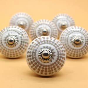 Puede incluir: Seis pomos de armario de cerámica blanca con un patrón de puntos dorados y detalles plateados. Los pomos están dispuestos en semicírculo.