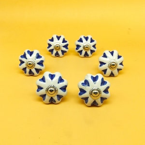 May include: Six white ceramic knobs with blue and gold accents. The knobs are shaped like flowers with five petals and have a decorative design.