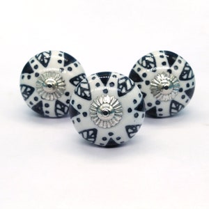 Op de afbeelding: Drie witte keramische knoppen met een geometrisch patroon in zwart en wit. De knoppen hebben een zilveren metalen midden.