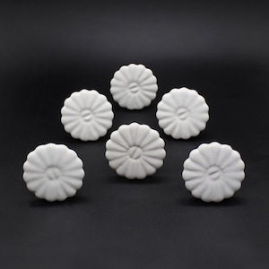 Op de afbeelding: Zes witte keramische ladegrepen met een bloemachtig ontwerp. De grepen hebben een verhoogd midden en een gegolfde rand.