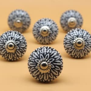 May include: Six white ceramic knobs with a black floral design and silver accents. The knobs are arranged in a circle on a light brown surface.