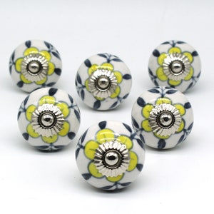 Op de afbeelding: Zes witte keramische knoppen met een geel bloemenontwerp en een zilveren midden. De knoppen hebben een blauw bloemenpatroon rond de randen.
