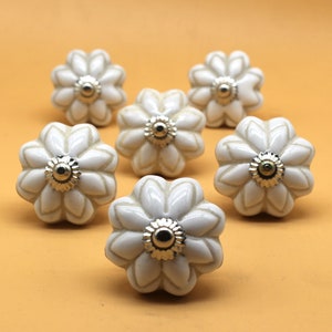 Op de afbeelding: Zeven witte keramische bloemvormige kastknoppen met zilverkleurige metalen accenten. De knoppen zijn gerangschikt op een oranje achtergrond. Elke knop heeft een glanzende afwerking en een gedetailleerd bloemblaadjesontwerp, geschikt voor meubels of decoratie.