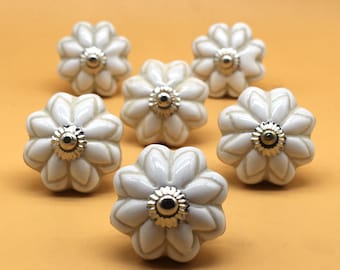 May include: Seven white ceramic flower-shaped cabinet knobs with silver-coloured metal accents. The knobs are arranged on an orange background. Each knob has a glossy finish and a detailed petal design, suitable for furniture or decor.