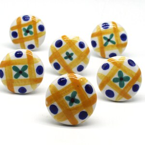 以下が含まれることがあります： 黄色と青色の幾何学模様が施されたセラミック製のキャビネットノブ6個。ノブは白地に黄色の格子模様と青いドットが施されています。各ノブの中央には緑色の花が描かれています。