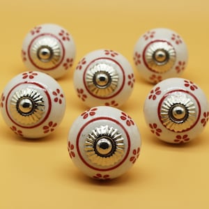 Op de afbeelding: Zes witte keramische kastknoppen met rode bloemenmotieven en zilveren accenten.