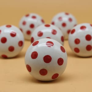 Könnte beinhalten: Sechs weiße Keramik-Kugeln mit roten Punkten. Die Kugeln sind in einem Halbkreis auf einer hellbraunen Oberfläche angeordnet.