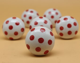 May include: Six white ceramic balls with red polka dots. The balls are arranged in a semi-circle on a light brown surface.