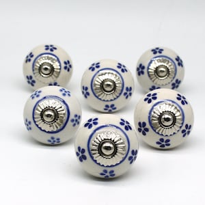 Pode incluir: Seis botões de cerâmica branca com desenhos florais azuis e detalhes prateados. Os botões são redondos e têm um centro elevado.