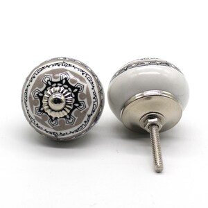 Op de afbeelding: Twee keramische kastknoppen met een witte achtergrond en een geometrisch ontwerp in grijs en zwart. De knop aan de linkerkant heeft een zilveren schroef, terwijl de knop aan de rechterkant een zilveren schroef en een zilveren basis heeft.