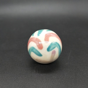 Op de afbeelding: Een witte keramische bol met een kleurrijk abstract ontwerp. De bol heeft roze en turquoise gebogen lijnen op een witte achtergrond.