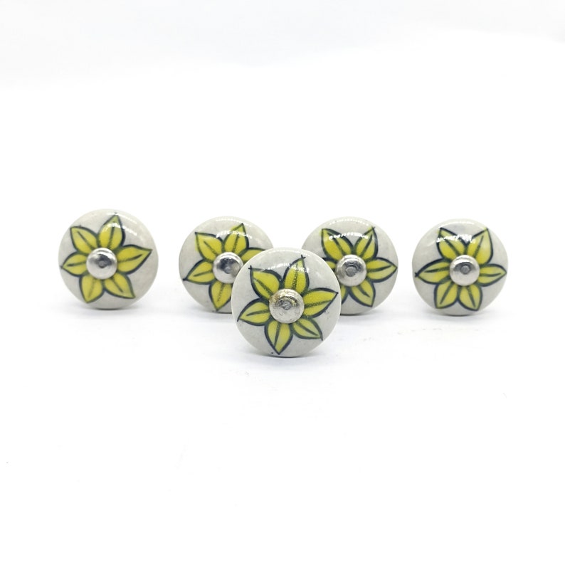K&ouml;nnte beinhalten: Vier Keramik-Schubladengriffe mit wei&szlig;em Hintergrund und gelbem Blumendesign. Die Blume hat einen dunkelgr&uuml;nen Umriss und eine silberne Mitte.